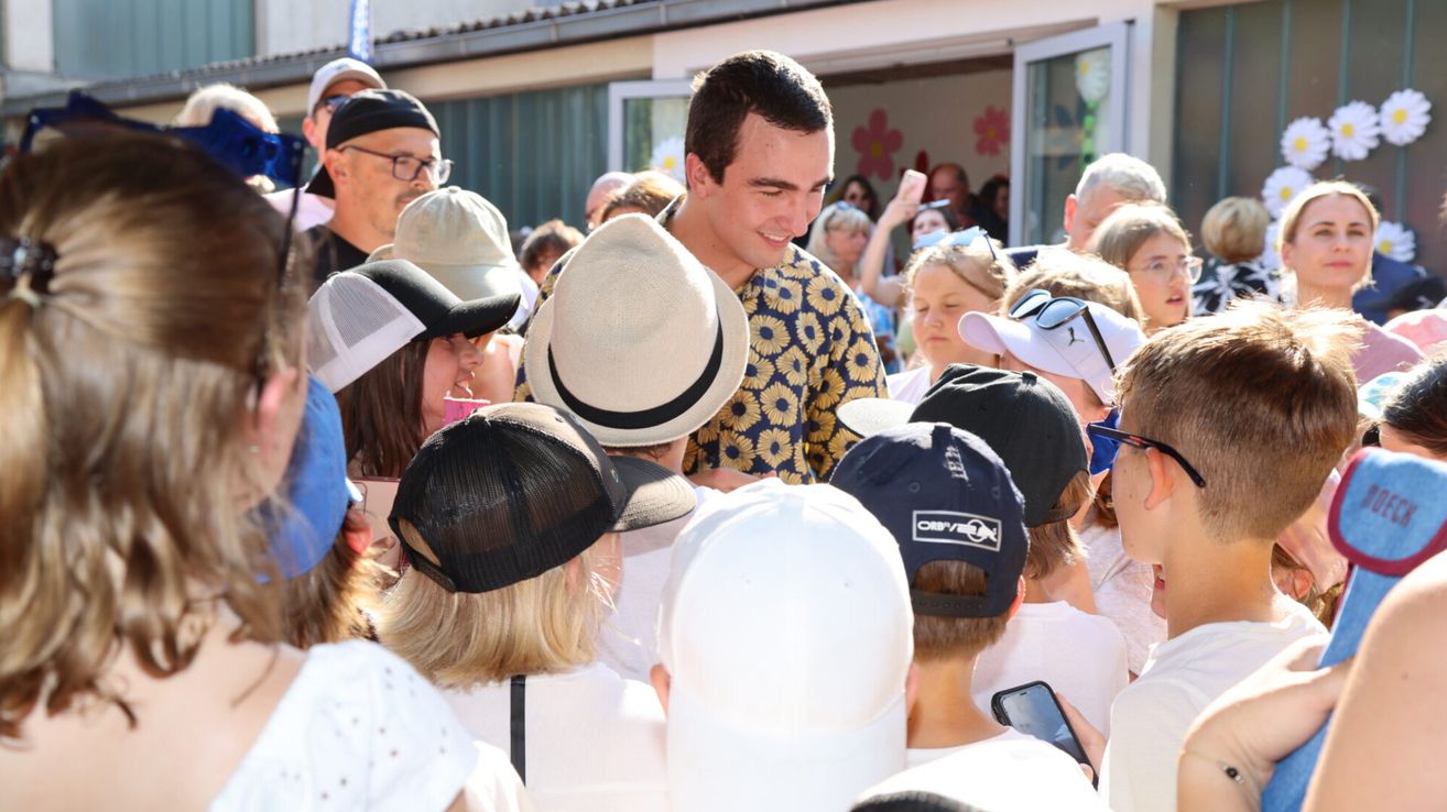 Ein Mann in einem Blumenhemd steht umgeben von Menschen, einige mit Hüten und Brillen, vor einem Gebäude. Leute machen Fotos und einer hält ein Handy.