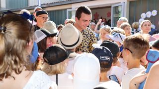 Ein Mann in einem Blumenhemd steht umgeben von Menschen, einige mit Hüten und Brillen, vor einem Gebäude. Leute machen Fotos und einer hält ein Handy.