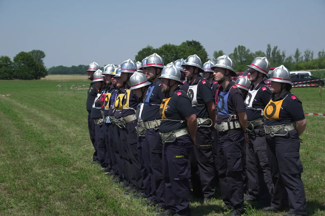 Eine Gruppe von Feuerwehrleuten in Uniform steht in einer Reihe auf einem Grasfeld. Sie tragen Helme mit Nummern, Gürtel und reflektierende Westen. Der Himmel ist klar, und Bäume sind in der Ferne.