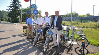 Vier Männer stehen neben Fahrrädern im Freien. Einer trägt Hosenträger, ein anderer trägt eine Brille. Sie befinden sich in der Nähe eines Fahrradstationsschildes.