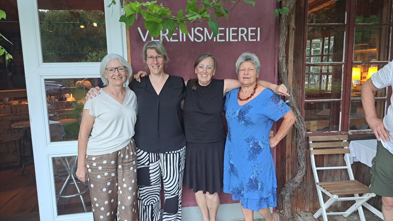 Vier Frauen in verschiedenen Kleidern stehen lächelnd vor einem Gebäude mit der Aufschrift 'Vereinsmeierei'. Sie tragen alle eine Brille.