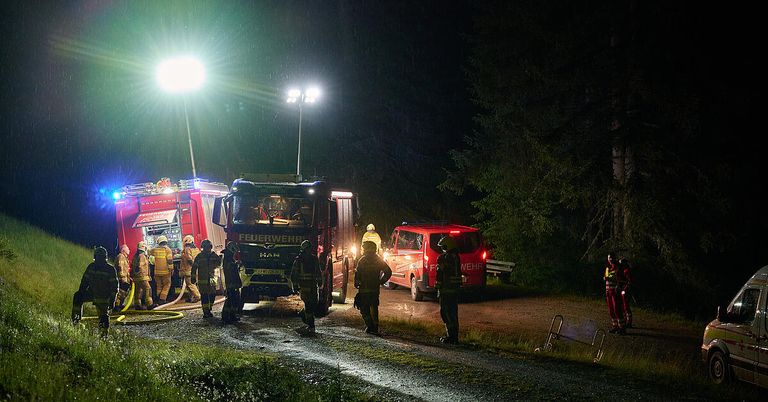 Feuerwehrleute kümmern sich um einen geparkten Feuerwehrwagen auf einer nassen Straße. Ein roter Van ist in der Nähe geparkt, und es stehen Leute um die Szene.