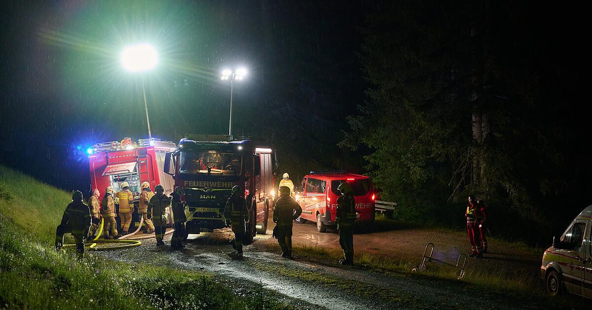 Feuerwehrleute kümmern sich um einen geparkten Feuerwehrwagen auf einer nassen Straße. Ein roter Van ist in der Nähe geparkt, und es stehen Leute um die Szene.