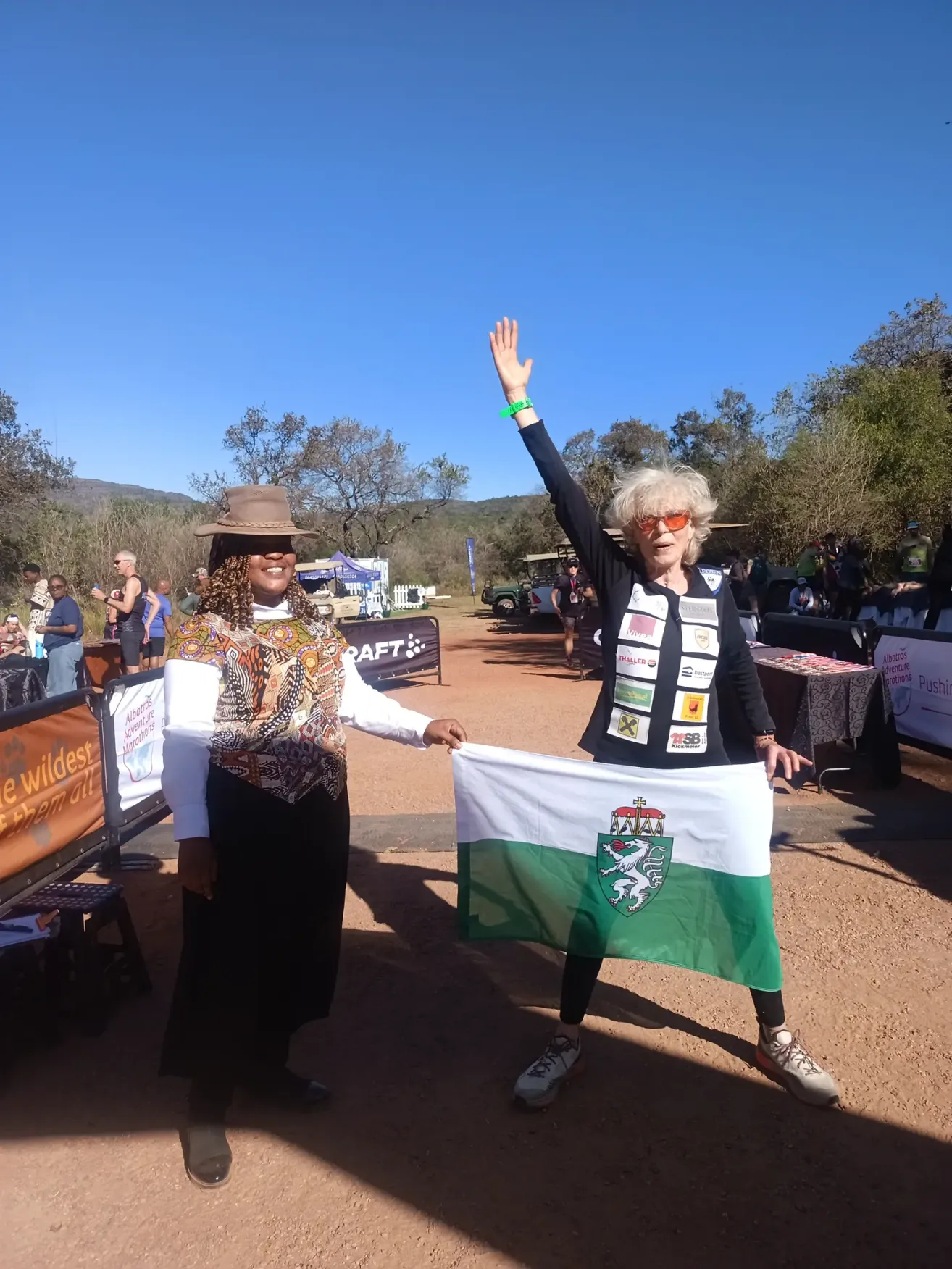 Zwei Frauen stehen auf einem Feld und schwenken eine Flagge. Eine trägt einen Hut und die andere ein grünes Armband. Ein Banner und Leute sind im Hintergrund.