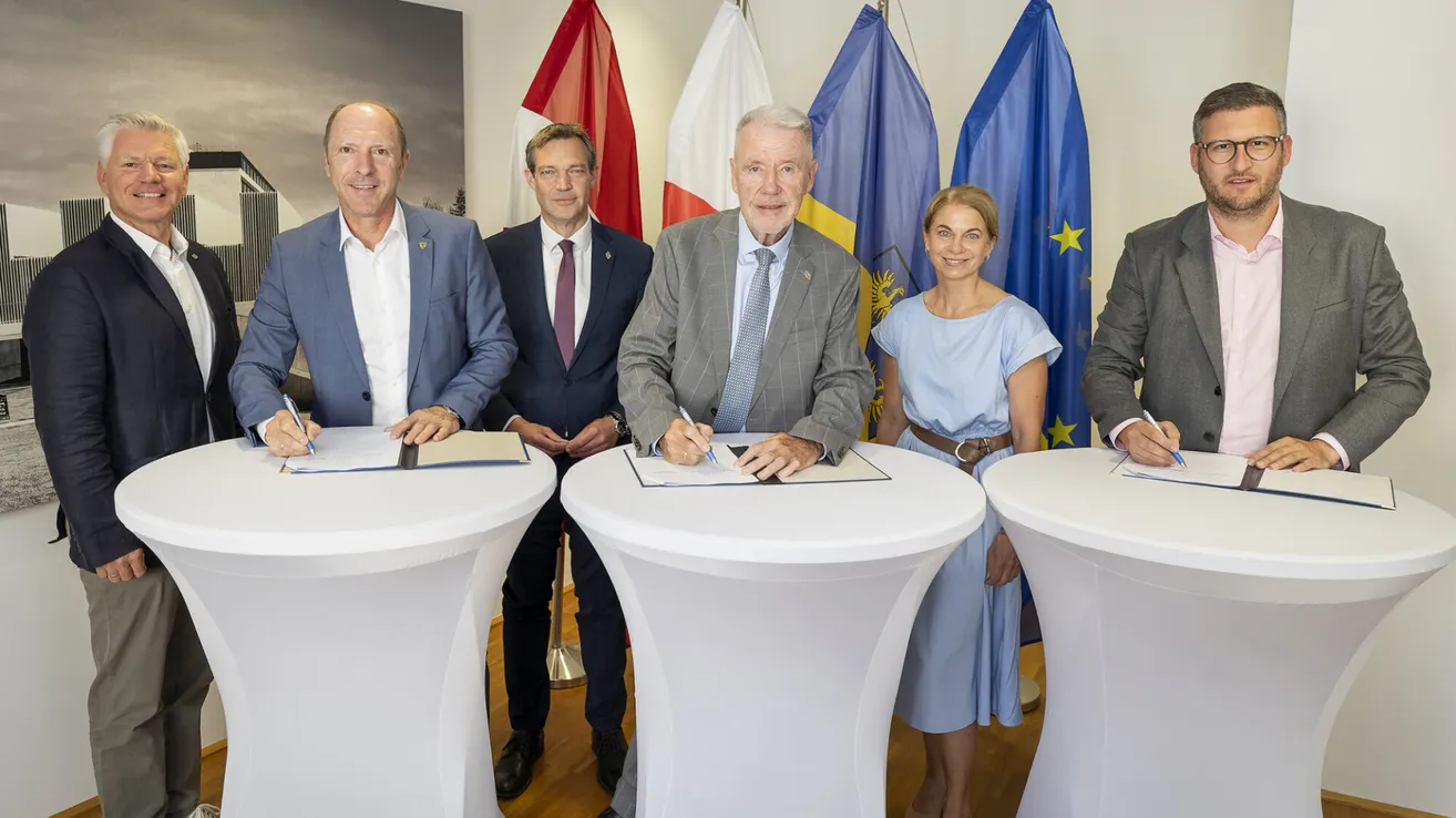 Fünf Personen posieren für ein Foto vor einem Tisch, wobei zwei von ihnen Papiere unterzeichnen. Sie sind formell gekleidet, mit Fahnen im Hintergrund.