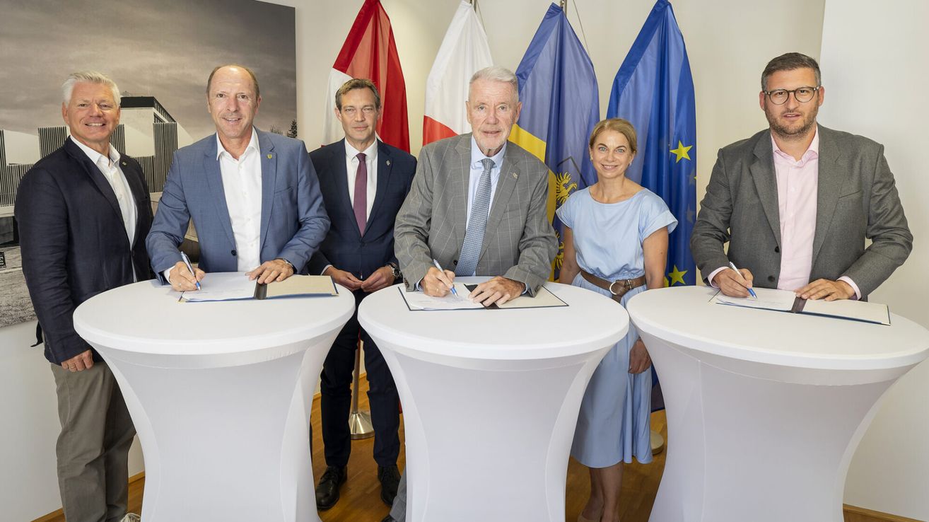 Fünf Personen posieren für ein Foto vor einem Tisch, wobei zwei von ihnen Papiere unterzeichnen. Sie sind formell gekleidet, mit Fahnen im Hintergrund.
