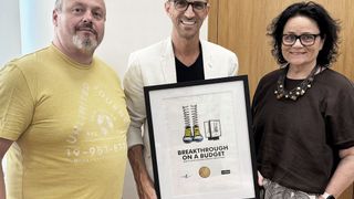 Drei Personen stehen vor einer Tür. Ein Mann hält ein gerahmtes Poster mit der Aufschrift Breakthrough on a Budget. Der Mann links trägt ein gelbes T-Shirt mit weißem Text und Zahlen.