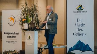Ein älterer Mann steht an einem Rednerpult mit Laptop und Mikrofon, möglicherweise gibt er eine Präsentation. Er trägt einen Anzug und eine Brille. Hinter ihm steht eine Pflanze mit Blumen und ein Banner mit Text und Logo.