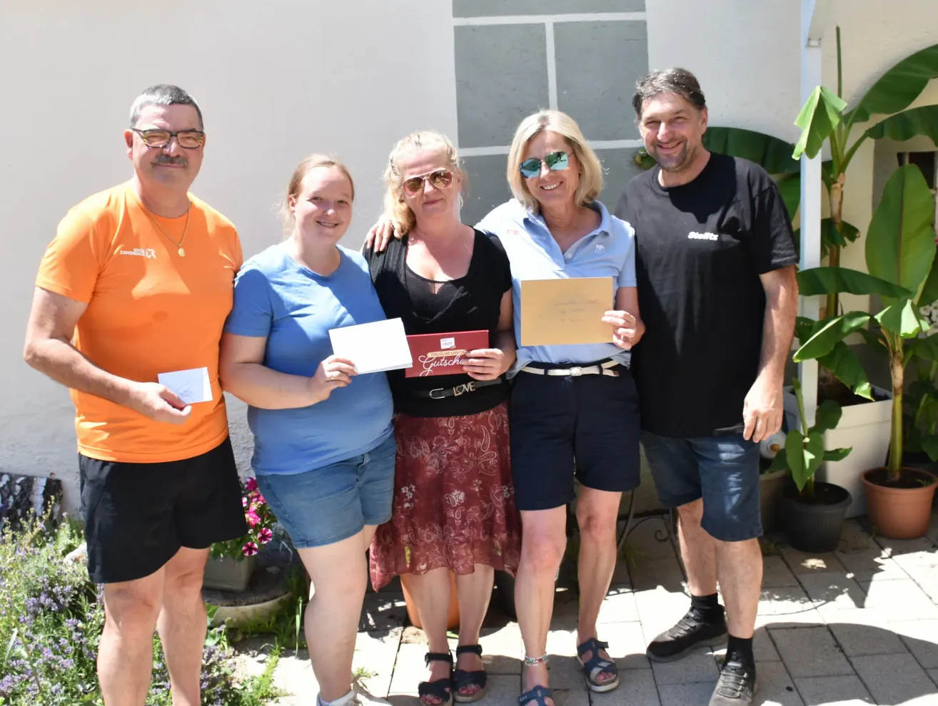Fünf Menschen stehen gemeinsam auf einer Terrasse und lächeln. Zwei Frauen und drei Männer halten Urkunden und eine rote Box in der Hand, wahrscheinlich zur Feier einer Leistung. Der Mann auf der linken Seite trägt ein orangefarbenes T-Shirt und die Frau auf der rechten Seite ein schwarzes T-Shirt. Hinter ihnen befindet sich eine weiße Wand.