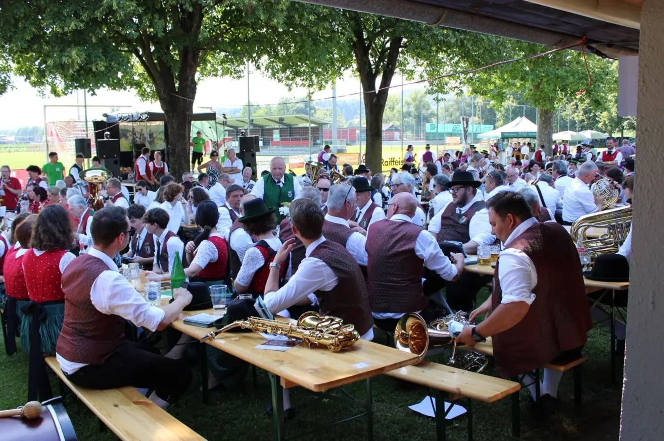 Eine große Gruppe von Menschen versammelt sich an einem Ort im Freien, möglicherweise für ein Musikfestival. Sie sitzen an Tischen mit Musikinstrumenten und Getränken. Bäume und ein Zelt sind im Hintergrund.