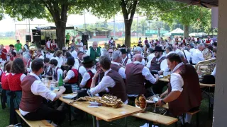 Eine große Gruppe von Menschen versammelt sich an einem Ort im Freien, möglicherweise für ein Musikfestival. Sie sitzen an Tischen mit Musikinstrumenten und Getränken. Bäume und ein Zelt sind im Hintergrund.