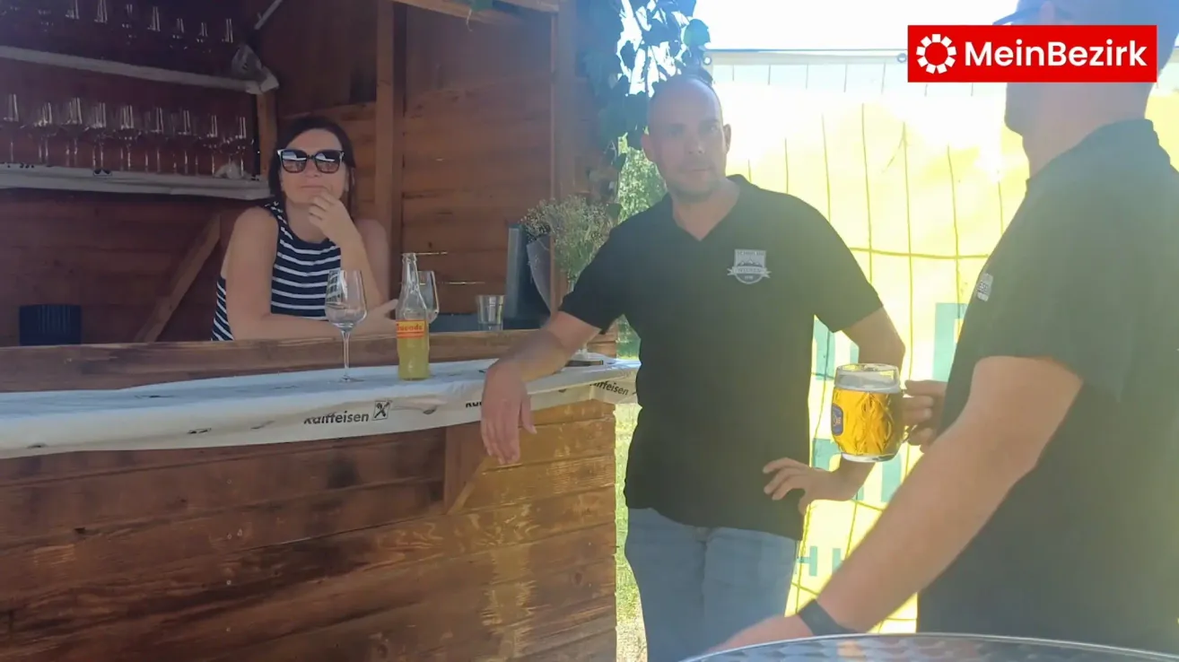 Ein Mann hält ein Bier in einer Bar. Eine Frau sitzt hinter der Theke, trägt eine Sonnenbrille und ein gestreiftes Shirt. Auf der Theke befinden sich eine Flasche und Gläser.
