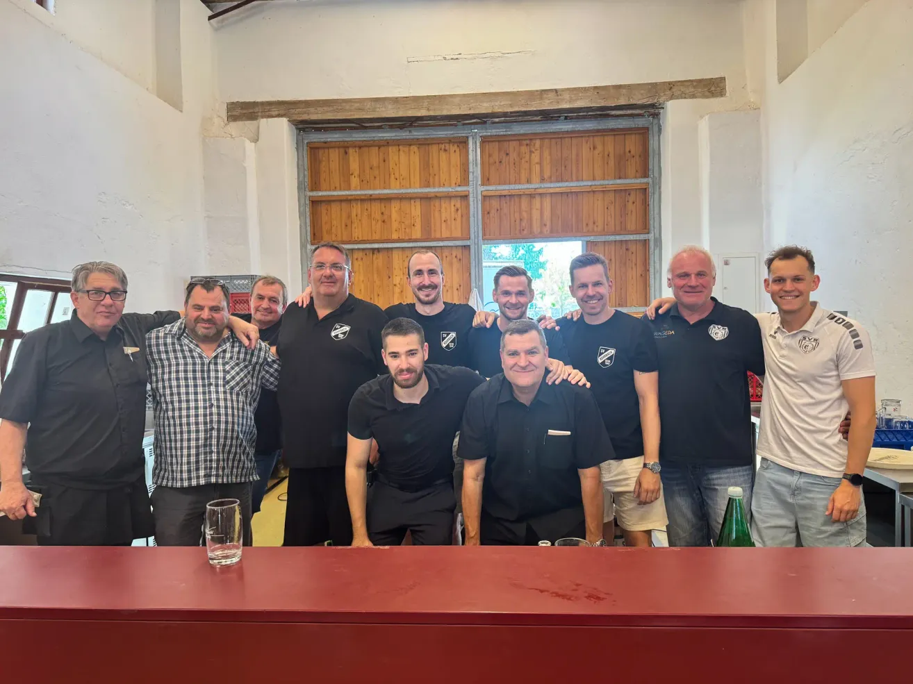 Eine Gruppe von Männern in schwarzen Shirts posiert für ein Foto in einem rustikalen Raum mit Holztüren. Ein Tisch mit einem Glas und einer grünen Flasche steht vor ihnen.