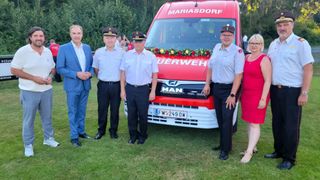 Vier Personen stehen vor einem roten Krankenwagen mit Blumen. Sie sind in Uniform. Hinter ihnen versammeln sich Leute, und Bäume sind sichtbar.