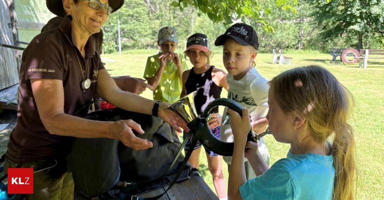 Eine Erwachsene reicht einem kleinen Mädchen ein Horn. Hinter ihnen schauen vier Kinder zu. Sie befinden sich auf einem Rasen.