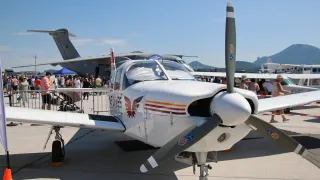 Ein weißes Flugzeug mit einem roten Logo ist auf dem Vorfeld geparkt, umgeben von einer Menschenmenge. Ein größeres Flugzeug ist im Hintergrund geparkt.
