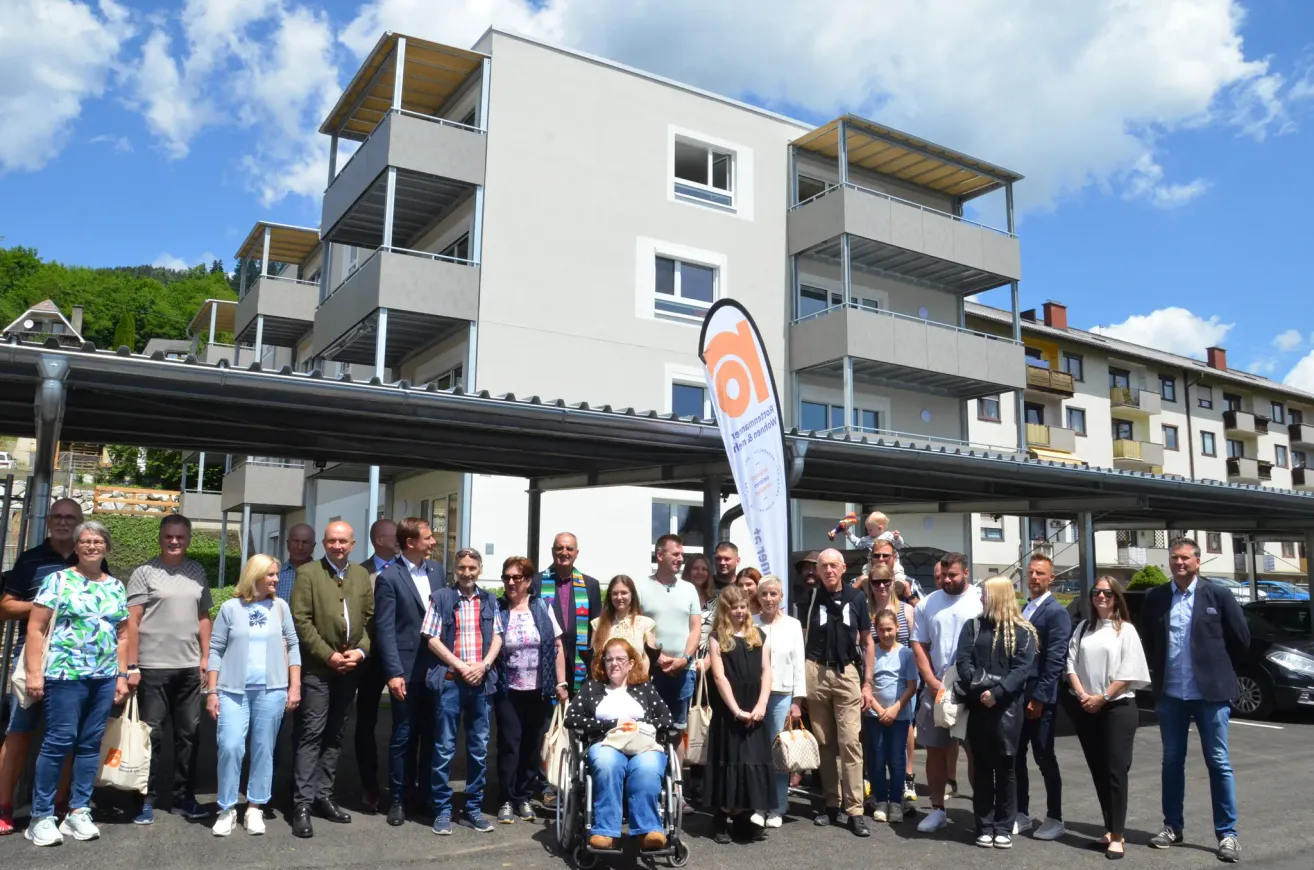 Eine Gruppe von Menschen posiert für ein Foto vor einem Gebäude mit mehreren Stockwerken und Balkonen. Der Himmel ist bewölkt.