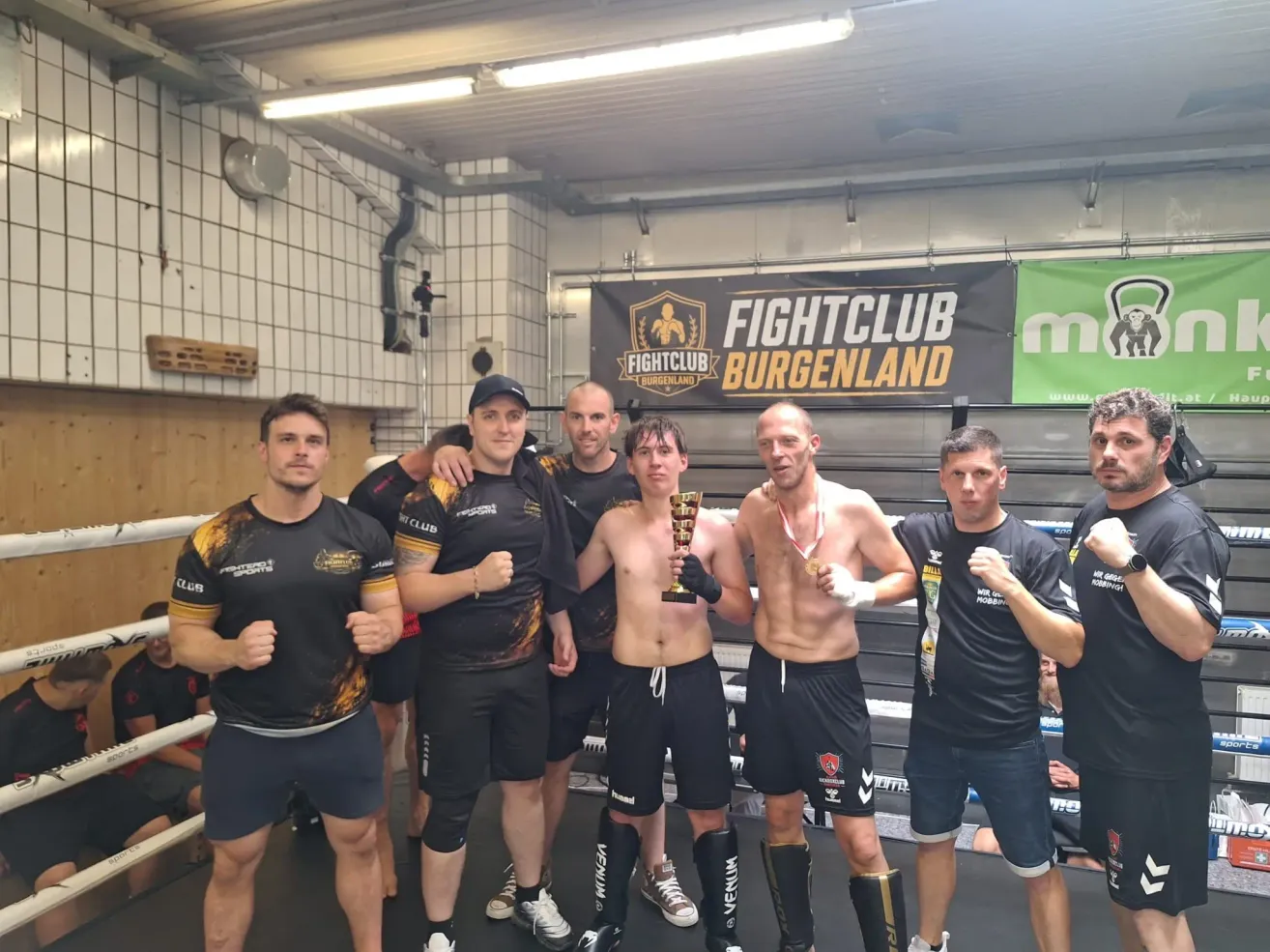 Sechs Männer in einem Fitnessstudio posieren für ein Foto. Ein Mann in der Mitte hält einen Pokal. Hinter ihnen ist ein Banner mit der Aufschrift 'FIGHTCLUB BURGENLAND'.