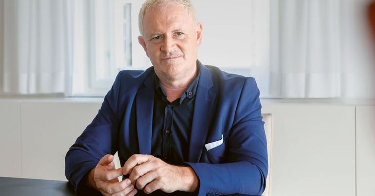 Ein Mann in einem blauen Anzug sitzt in einem Stuhl, Hände verschränkt, und lächelt leicht.