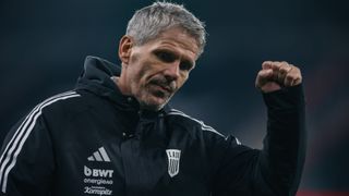 Ein Mann in einem schwarzen Jacke mit weißen Adidas Streifen und den Buchstaben LASK auf der linken Seite. Er sieht nach vorne und scheint zu schreien.