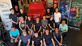 Eine Gruppe von Menschen, darunter Männer und Frauen, posiert für ein Foto vor einem roten Auto in einer Werkstatt. Sie tragen alle Freizeitkleidung.