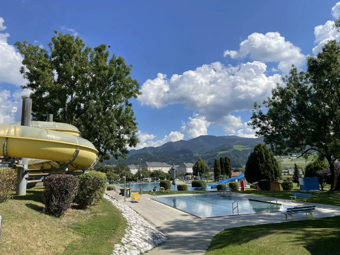 Ein Freibad mit einer Rutsche, Bäumen und Blick auf Berge. Der Himmel ist blau mit Wolken.