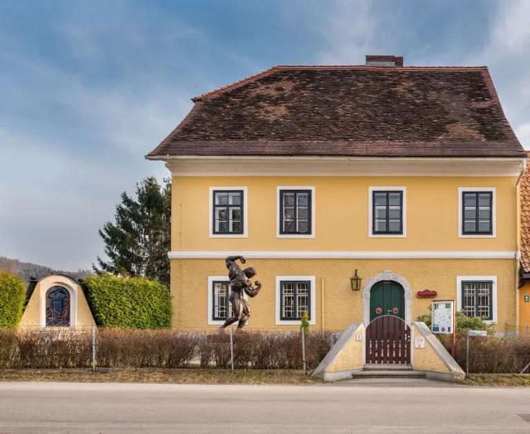 A yellow house with brown roof and multiple windows. A statue is in front of the house. The house has a brown gate.