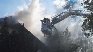Feuerwehrleute benutzen eine Kran, um auf das Dach eines brennenden Gebäudes zu gelangen. Rauch steigt aus dem Gebäude auf, und der Kran spritzt Wasser.