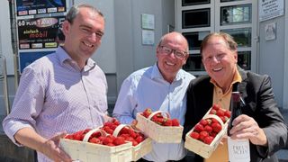 Drei Männer lächeln und halten Körbe mit Erdbeeren in den Händen. Sie stehen vor einem Gebäude.