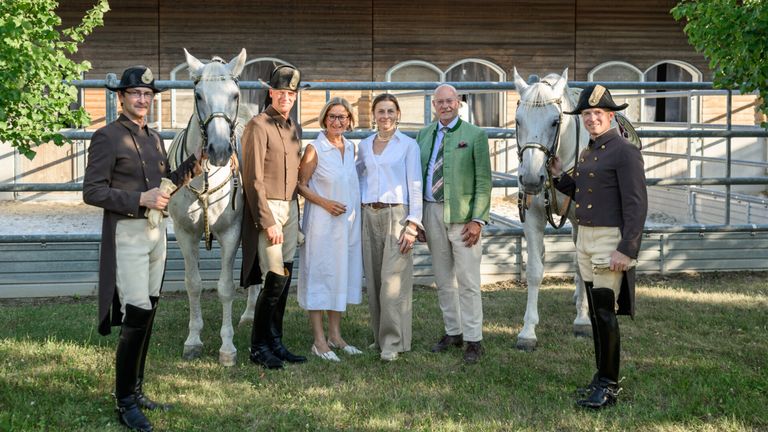 Five people in formal attire stand in front of two white horses. Two men wear hats and boots. Two women wear glasses and white blouses. A man in a green jacket stands between them.