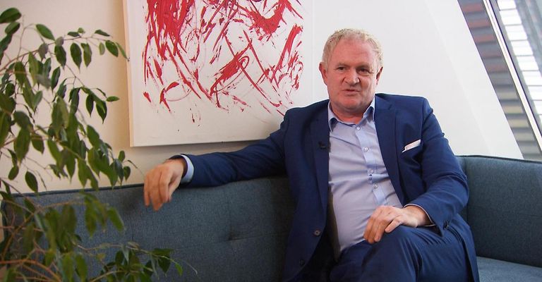 Ein Mann in einem blauen Anzug sitzt auf einer blauen Couch vor einem roten Gemälde.