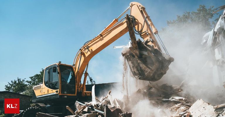A yellow excavator is demolishing debris in an outdoor setting, with dust rising from the operation.