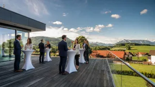 Eine Gruppe von Menschen in Anzügen unterhält sich auf einer Dachterrasse, umgeben von weißen Tischen und einem Glasgeländer, mit Bergen und einem roten Gebäude im Hintergrund.