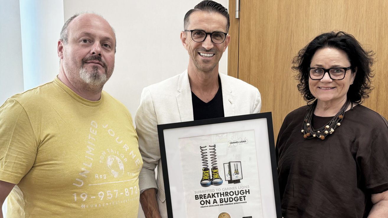 Drei Männer stehen eng beieinander. Der Mann in der Mitte hält einen gerahmten Preis und lächelt. Der Mann auf der linken Seite trägt ein gelbes T-Shirt mit Text und Zahlen.