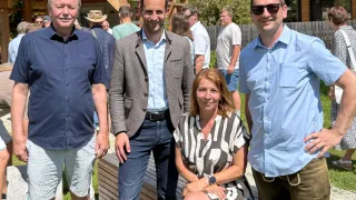 Vier Personen, drei Männer und eine Frau, posieren für ein Foto in einer Außenumgebung. Dahinter befindet sich ein Holzzaun und ein Haus.