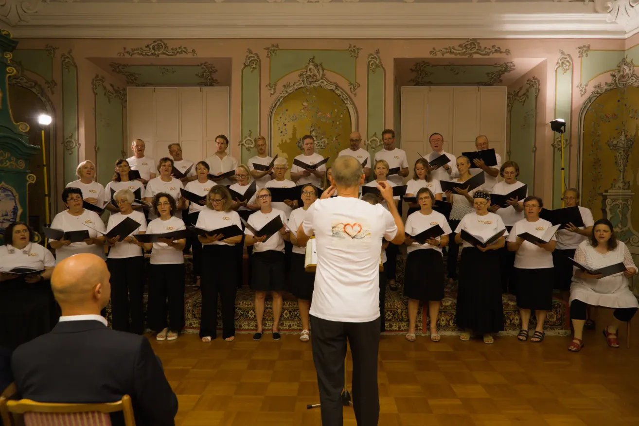 Ein Chor von Erwachsenen in Weiß und Schwarz steht in einem eleganten Raum, singt und wird von einem Mann in einem weißen T-Shirt dirigiert.