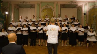 Ein Chor von Erwachsenen in Weiß und Schwarz steht in einem eleganten Raum, singt und wird von einem Mann in einem weißen T-Shirt dirigiert.
