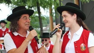 Ein Mann und eine Frau singen in Mikrofone. Die Frau lächelt und der Mann schaut sie an. Dahinter sind verschwommene Bilder von Menschen.