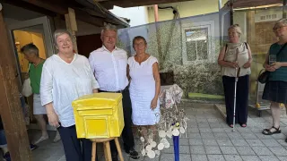 Drei Personen, zwei Frauen und ein Mann, stehen zusammen vor einer gelben Box auf einem Hocker. Hinter ihnen steht eine Frau mit einem Stock. Im Hintergrund befindet sich ein großes Gemälde eines Hauses und Bäume.