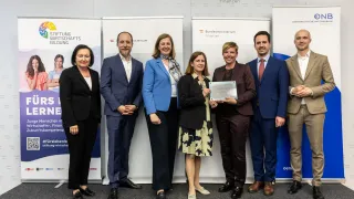 Eine Gruppe von Fachleuten in formeller Kleidung steht für ein Foto zusammen, einer hält ein Zertifikat. Dahinter werden Banner gezeigt, zwei davon tragen die Aufschrift 'Bundesministerium Finanzen'.
