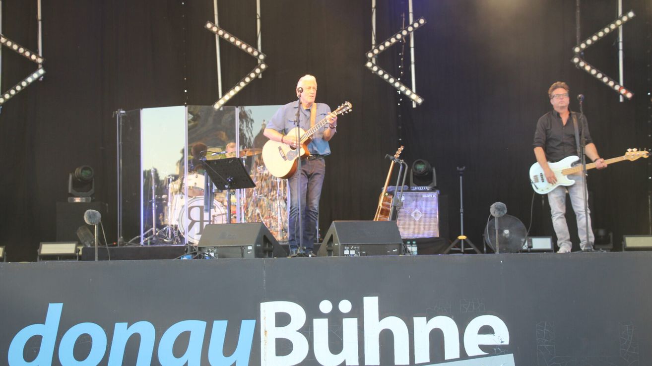 Ein Musiker spielt eine Gitarre auf der Bühne mit einem Schlagzeug im Hintergrund und Lautsprechern auf beiden Seiten.