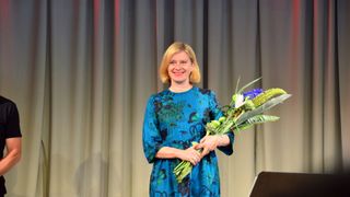 Eine Frau in einem blauen Kleid steht auf der Bühne, lächelt und hält einen Blumenstrauß vor einem grauen Vorhang.