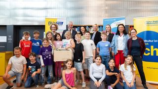 Eine Gruppe von Kindern und Erwachsenen posiert für ein Foto in einer Bibliothek. Einige Kinder halten eine Box mit Büchern. Hinter ihnen steht ein Banner mit der Aufschrift BHW Bildung ist Wert.