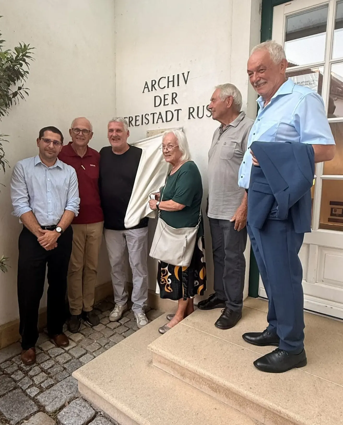 Sechs lächelnde Personen stehen vor einer Wand mit der Aufschrift 'Archiv der Widerstand Russ'. Sie tragen Freizeitkleidung. Eine Frau hält eine weiße Tasche.