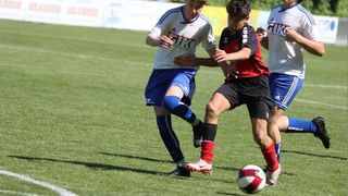Zwei Fußballspieler sind in einem Spiel auf einem Rasenfeld. Ein Spieler in Weiß versucht, den Ball zu kicken, während ein anderer in Rot versucht, ihn zu blocken.