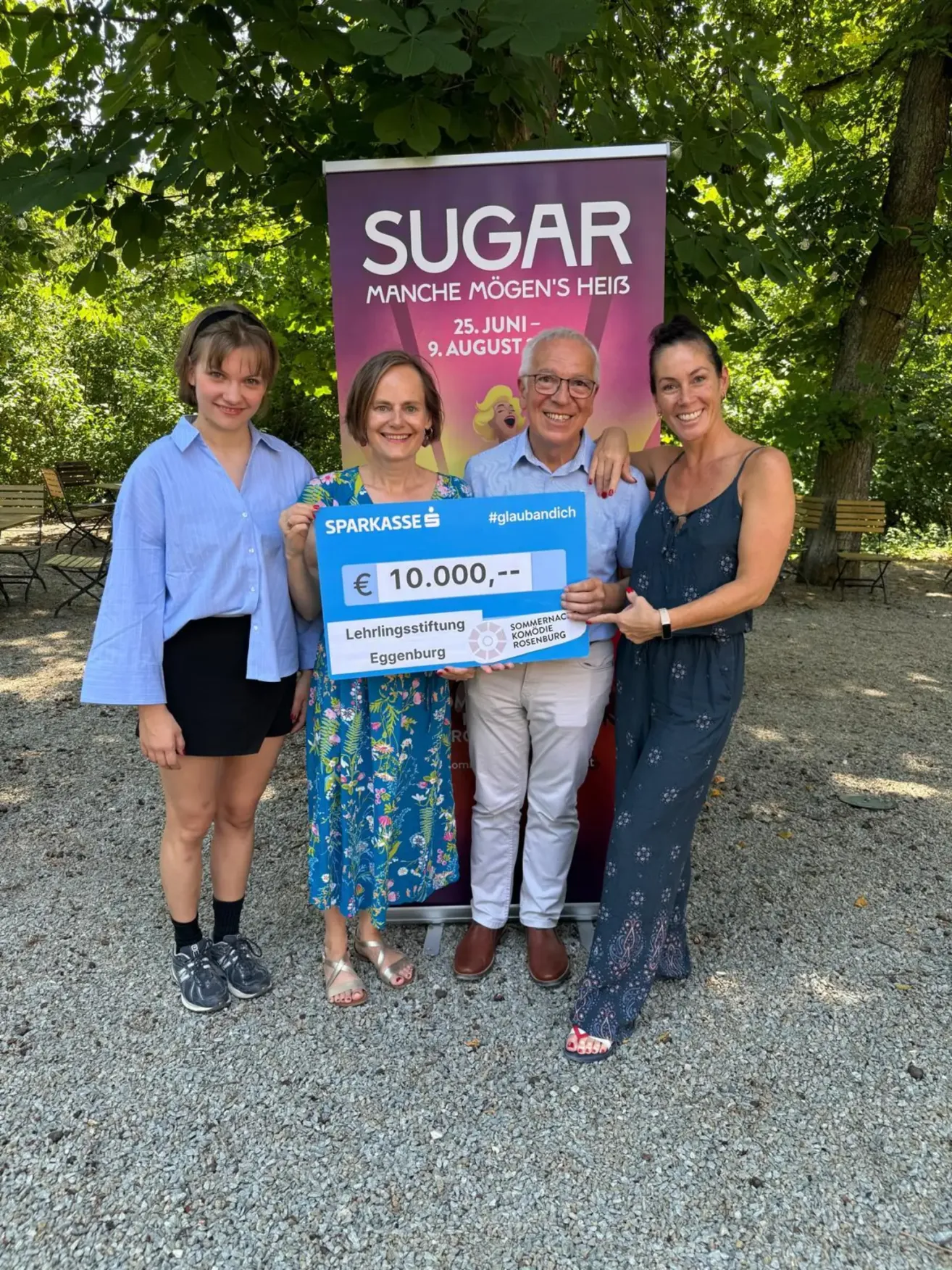 Drei Personen stehen vor einem Banner mit dem Namen Sugar. Sie halten einen Scheck über 10.000 Euro.