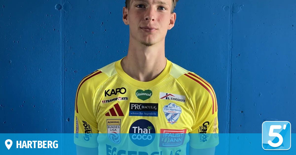 Ein Mann steht vor einer blauen Wand und trägt ein gelbes Trikot mit verschiedenen Sponsorenlogos. Er schaut geradeaus.