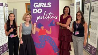 Drei Frauen stehen vor einem Banner mit der Aufschrift 'Girls Do Politics'. Sie lächeln und zeigen auf das Banner.