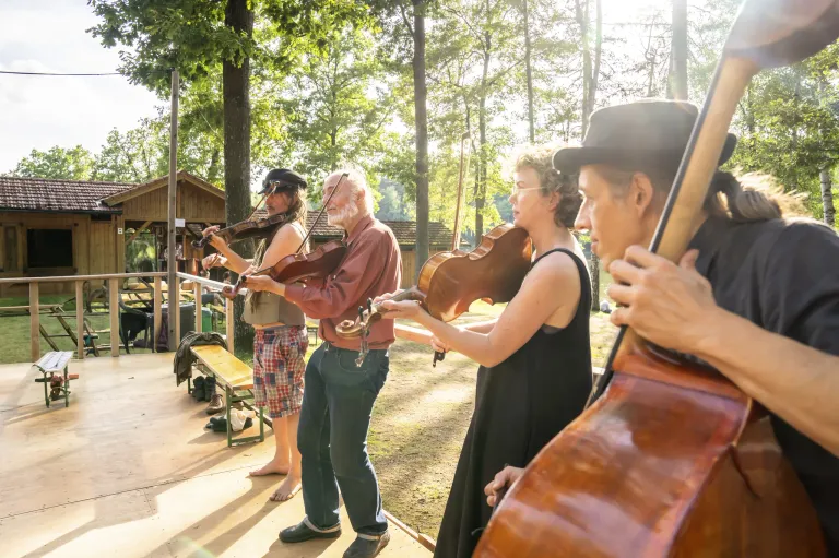 Ein Quartett von Musikern, jeder mit einer Geige, spielt auf einer Bühne, umgeben von Bäumen. Ein Geiger trägt einen Hut, eine Geigerin Brille. Die Bühne befindet sich in einem bewaldeten Bereich mit einer hölzernen Bank in der Nähe.