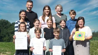 Eine Gruppe lächelnder Erwachsener und Kinder steht zusammen, hält Urkunden und einen gelben Pokal. Einige tragen eine Brille, und eine Frau hält ein gelbes Papier mit einem Design.